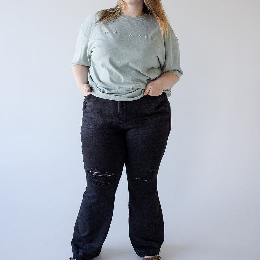 A person stands against a plain backdrop, wearing a light green T-shirt paired with high-waisted black jeans and sandals, hands casually tucked in pockets.