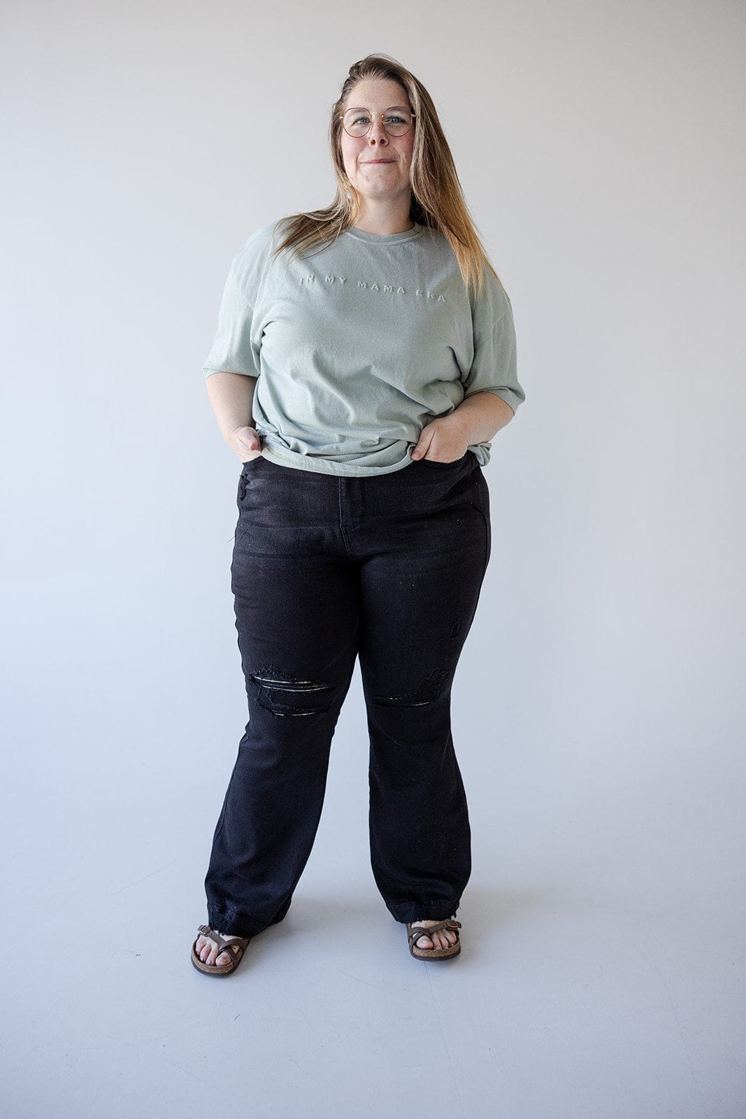 A person stands against a plain backdrop, wearing a light green T-shirt paired with high-waisted black jeans and sandals, hands casually tucked in pockets.