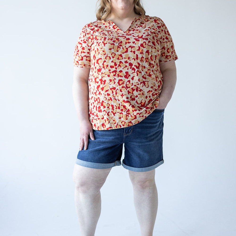 A woman with medium-length hair wearing a patterned blouse and a pair of Judy Blue Tummy Control Shorts with Cooling Technology, along with sandals, stands against a plain white background.