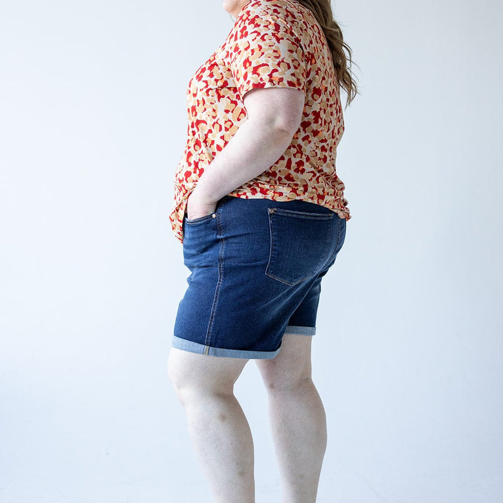 A person stands in profile against a plain background, wearing a red floral top, Judy Blue Tummy Control Shorts with Cooling Technology by Judy Blue, and sandals. They have their hands in their pockets.