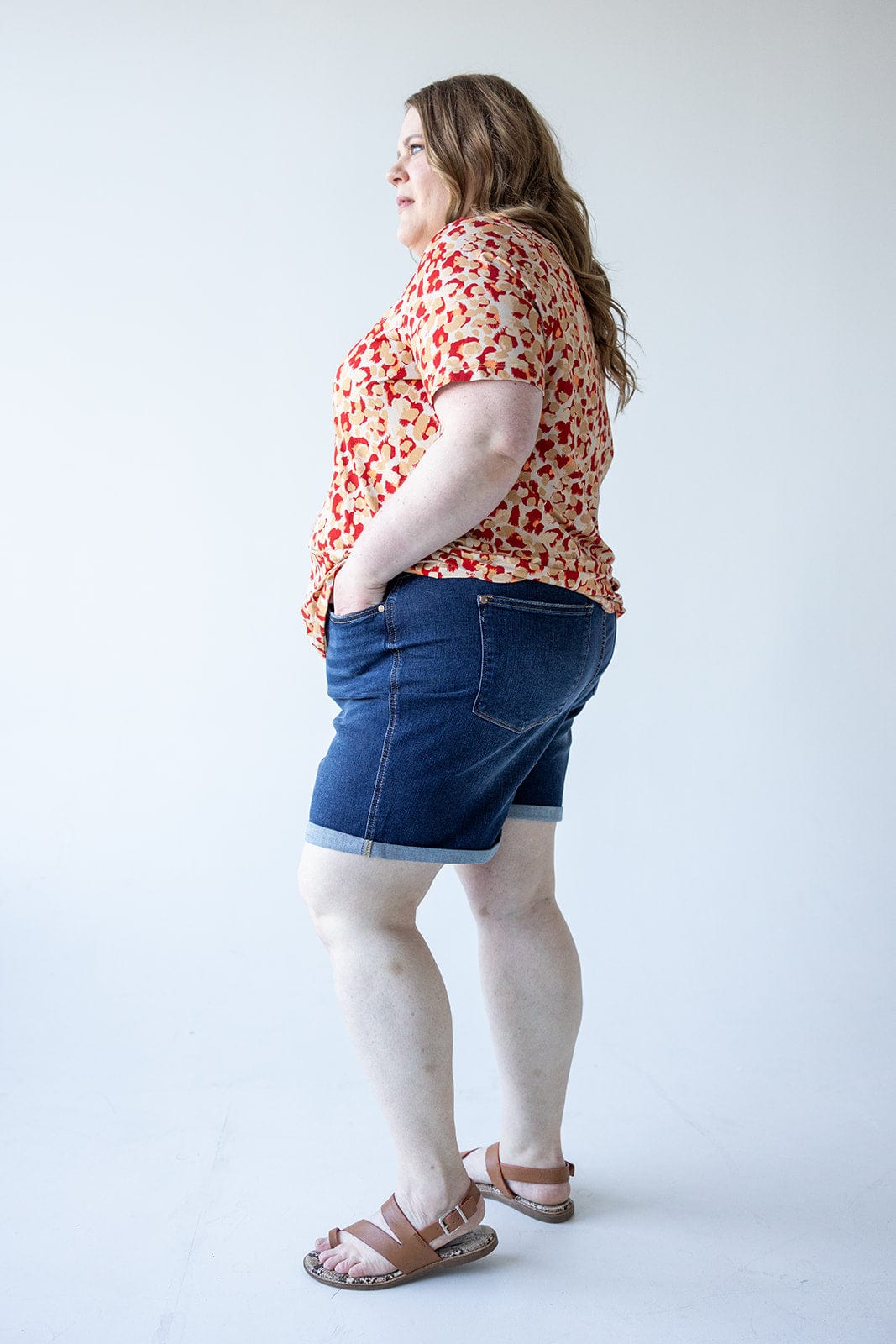 A person stands in profile against a plain background, wearing a red floral top, Judy Blue Tummy Control Shorts with Cooling Technology by Judy Blue, and sandals. They have their hands in their pockets.