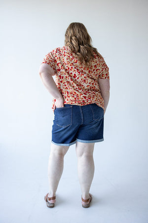 A person wearing a patterned shirt, Judy Blue Tummy Control Shorts with Cooling Technology, and sandals stands with their back turned in a white studio setting.