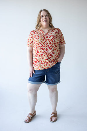 A woman with medium-length hair wearing a patterned blouse and a pair of Judy Blue Tummy Control Shorts with Cooling Technology, along with sandals, stands against a plain white background.