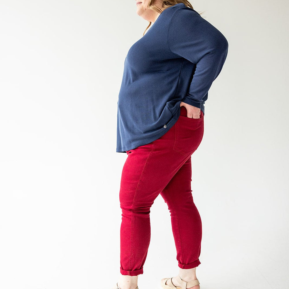 A person wearing a blue long-sleeve shirt, Judy Blue Tummy Control Skinny Jeans in Scarlet, and beige sandals stands with hands on hips against a plain background.