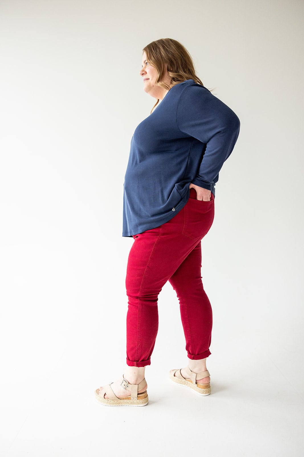 A person wearing a blue long-sleeve shirt, Judy Blue Tummy Control Skinny Jeans in Scarlet, and beige sandals stands with hands on hips against a plain background.