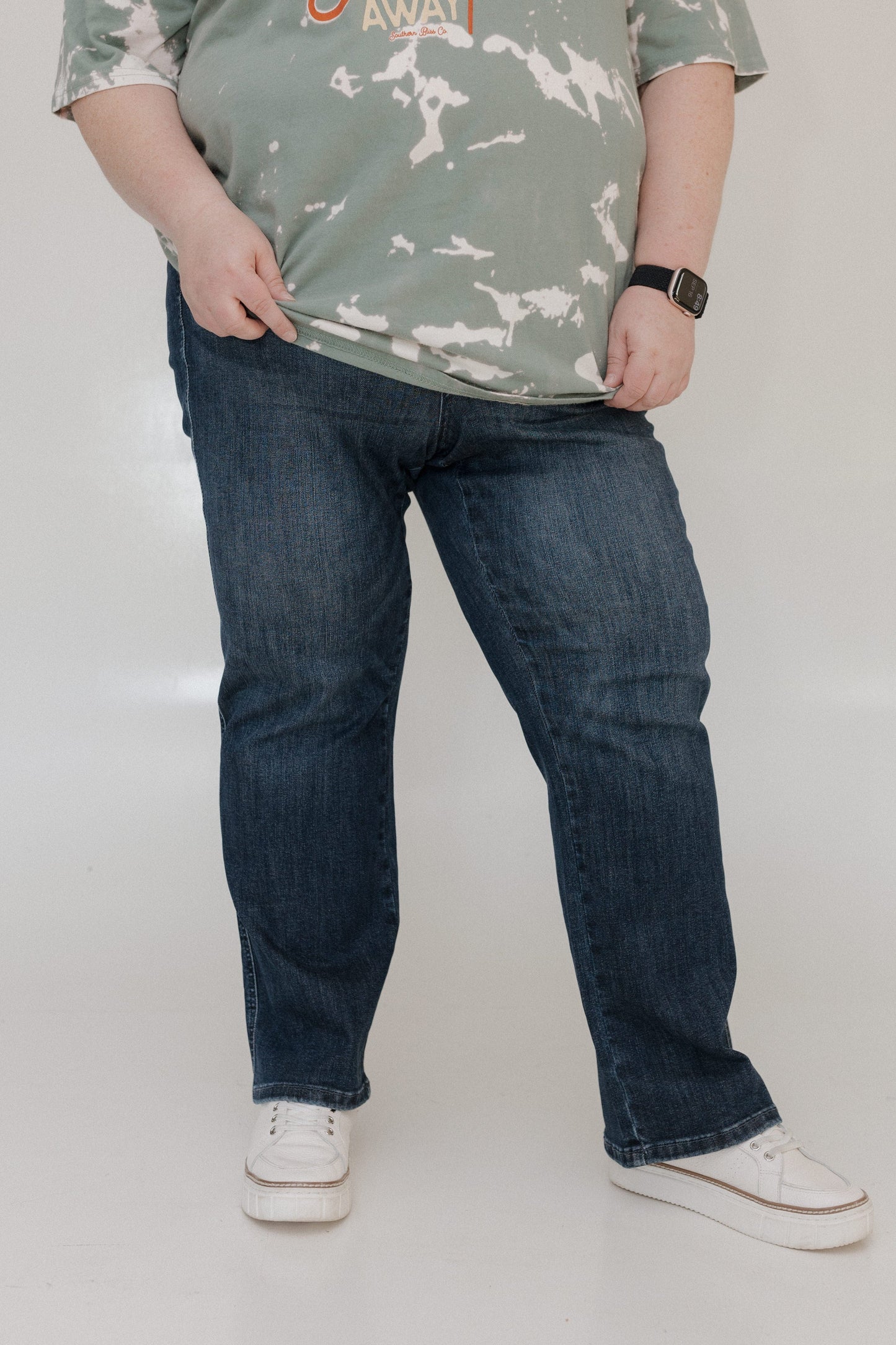 Person wearing a camouflage shirt, blue jeans, and white sneakers on a plain background