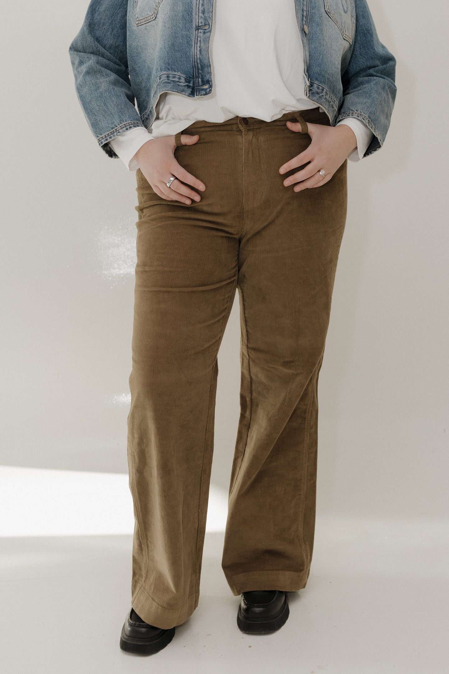 Person wearing brown corduroy pants with a denim jacket and white shirt on a plain background