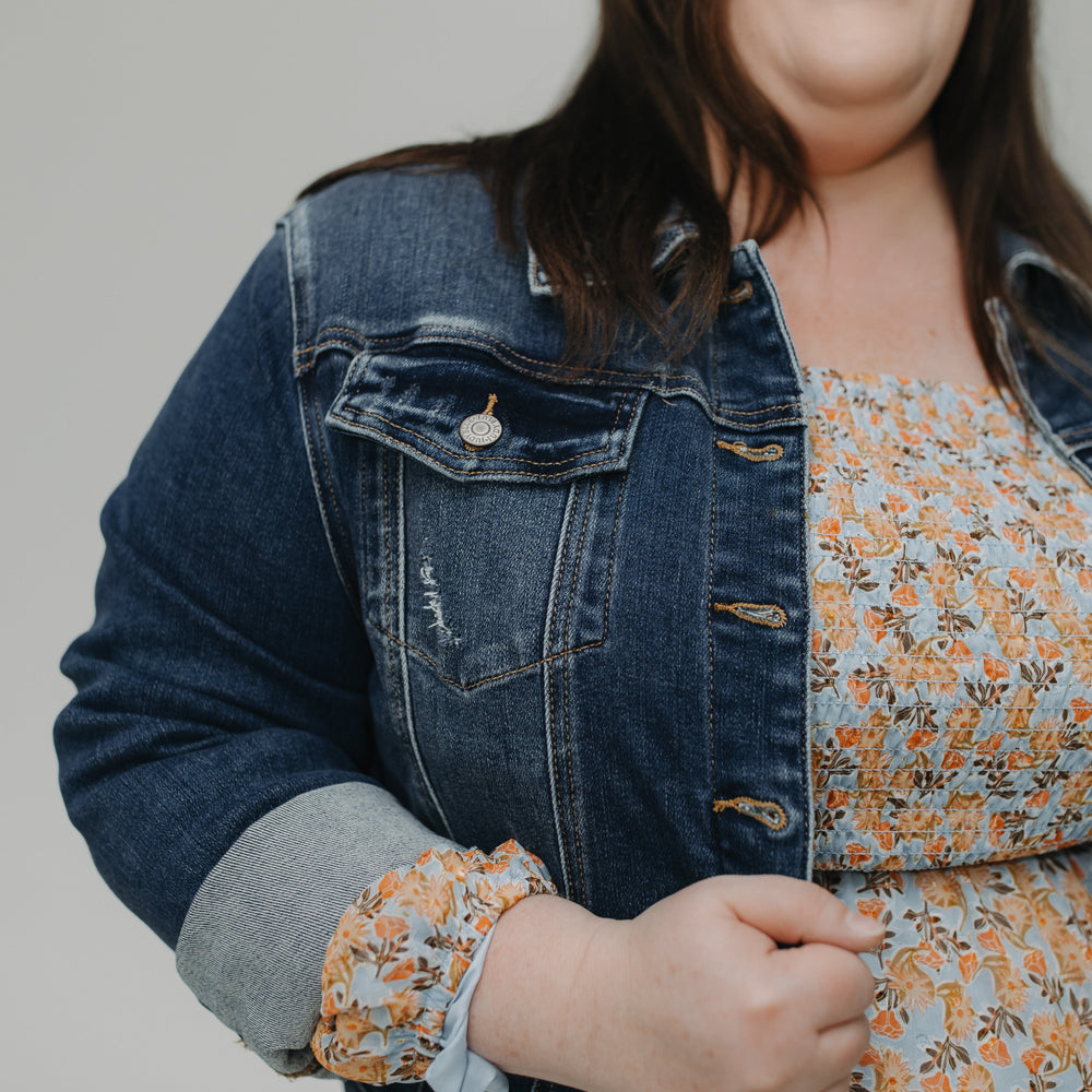 Woman wearing a denim jacket over a floral dress against a plain background