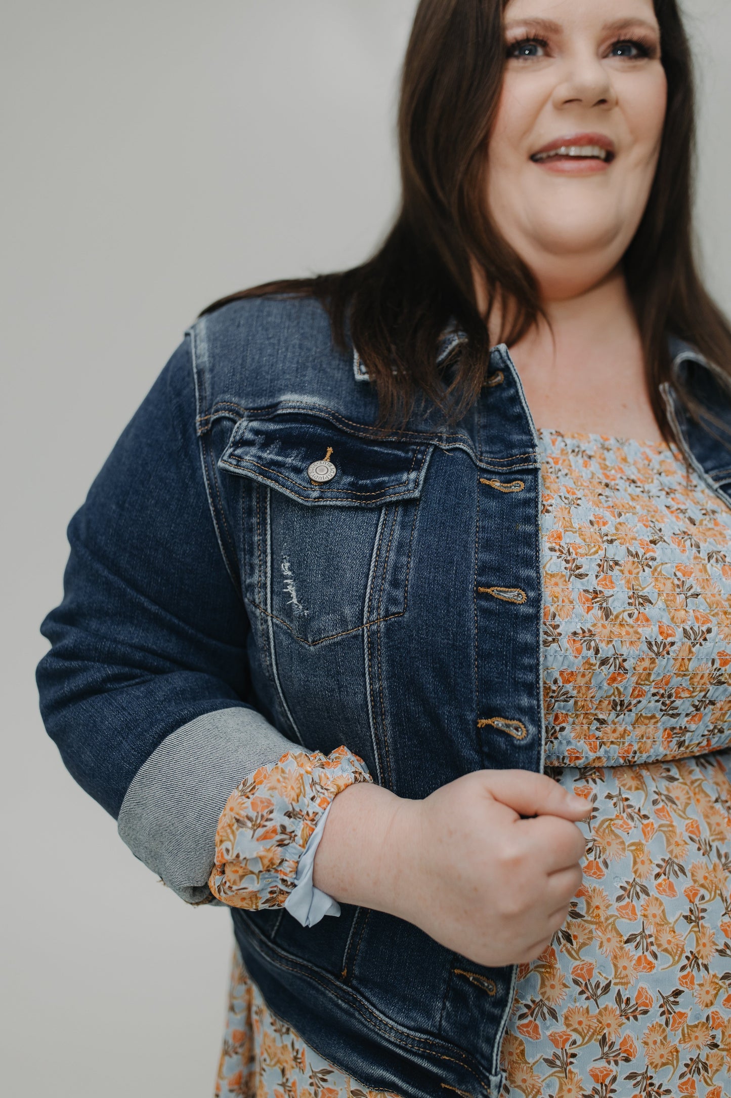 Woman wearing a denim jacket over a floral dress against a plain background