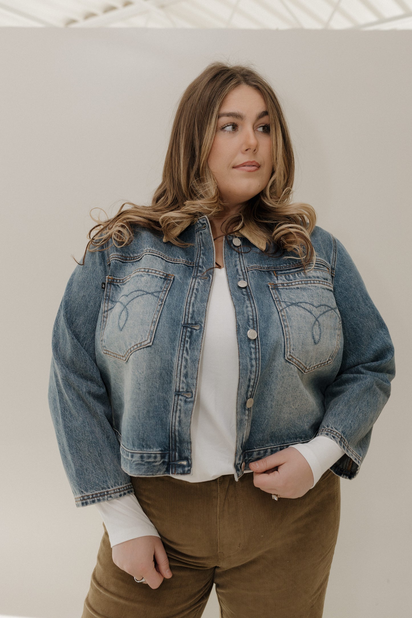 Woman wearing a denim jacket over a white shirt and brown pants against a plain background