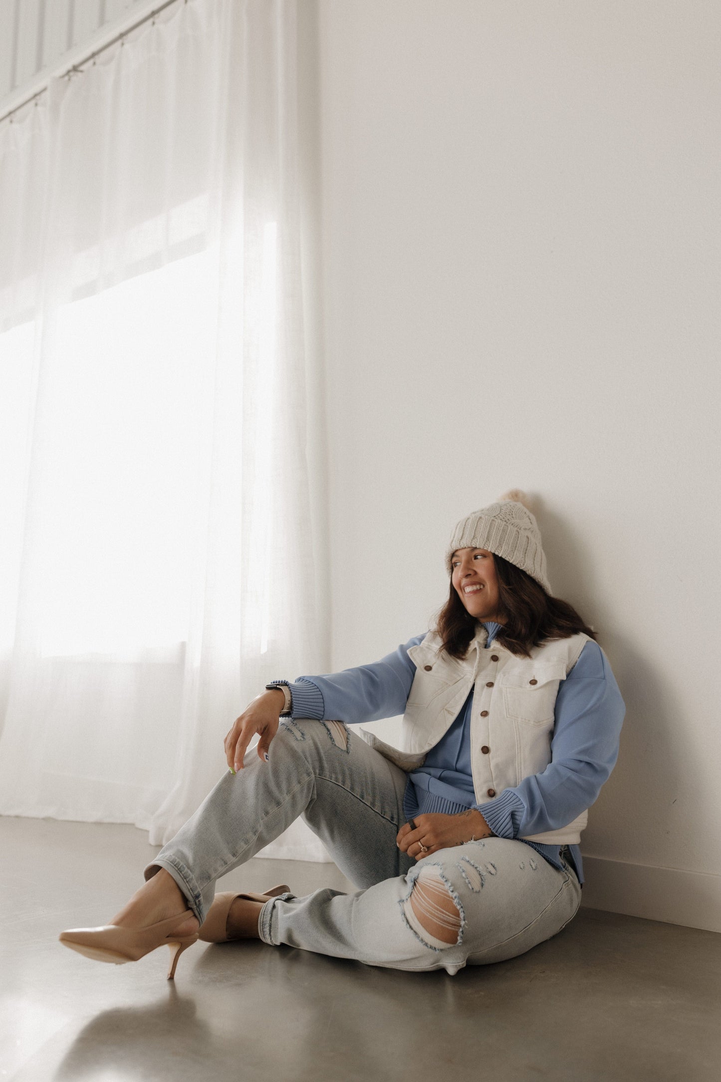 Person sitting on the floor against a white wall wearing a light blue shirt, beige vest, gray pants, and a beanie.