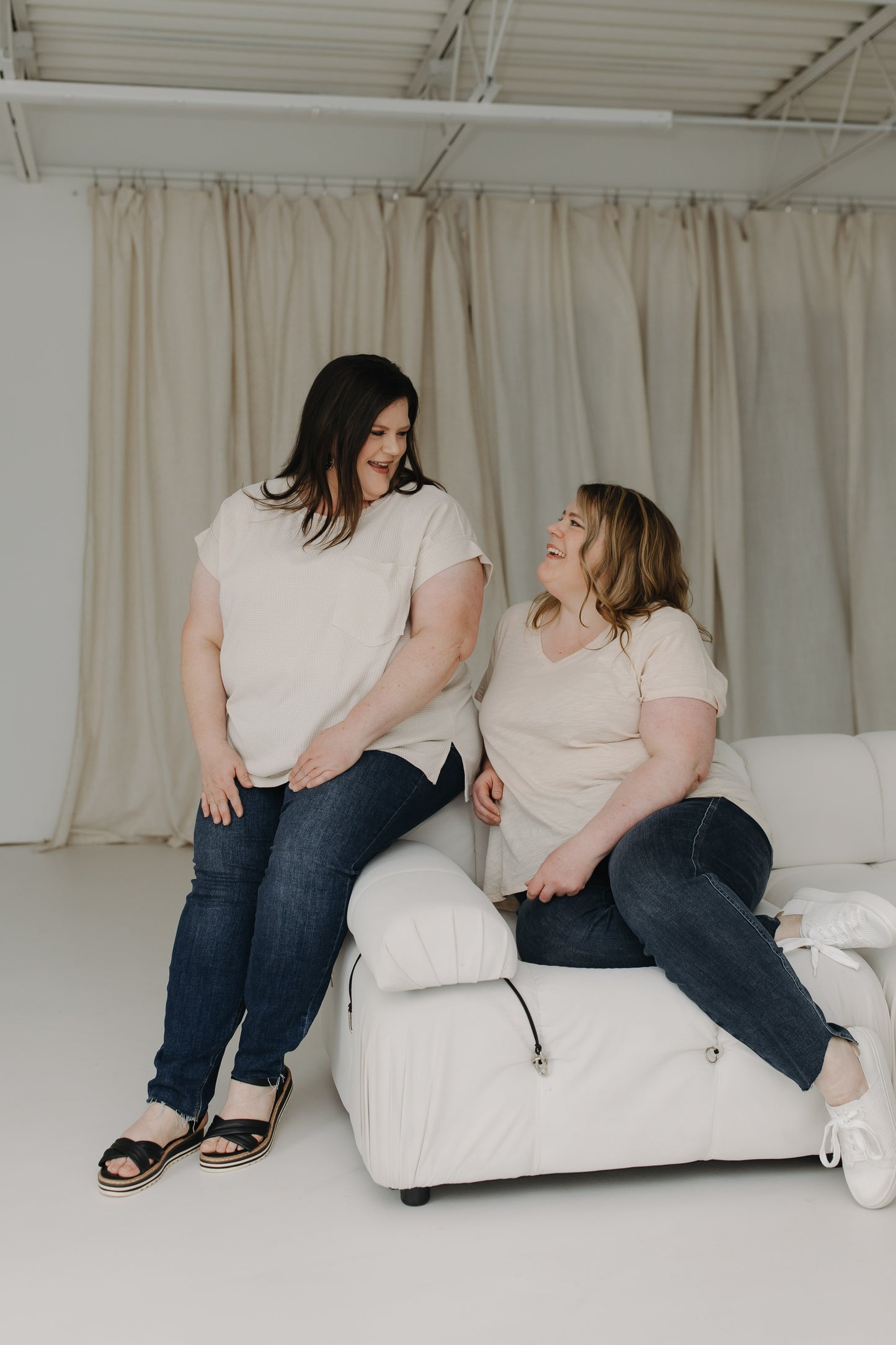 Two women sitting on a white couch in a room with beige curtains.