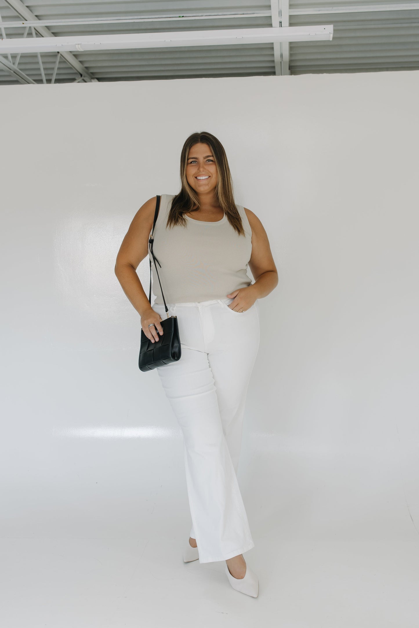 Woman wearing a beige top and white pants, holding a black handbag against a plain background