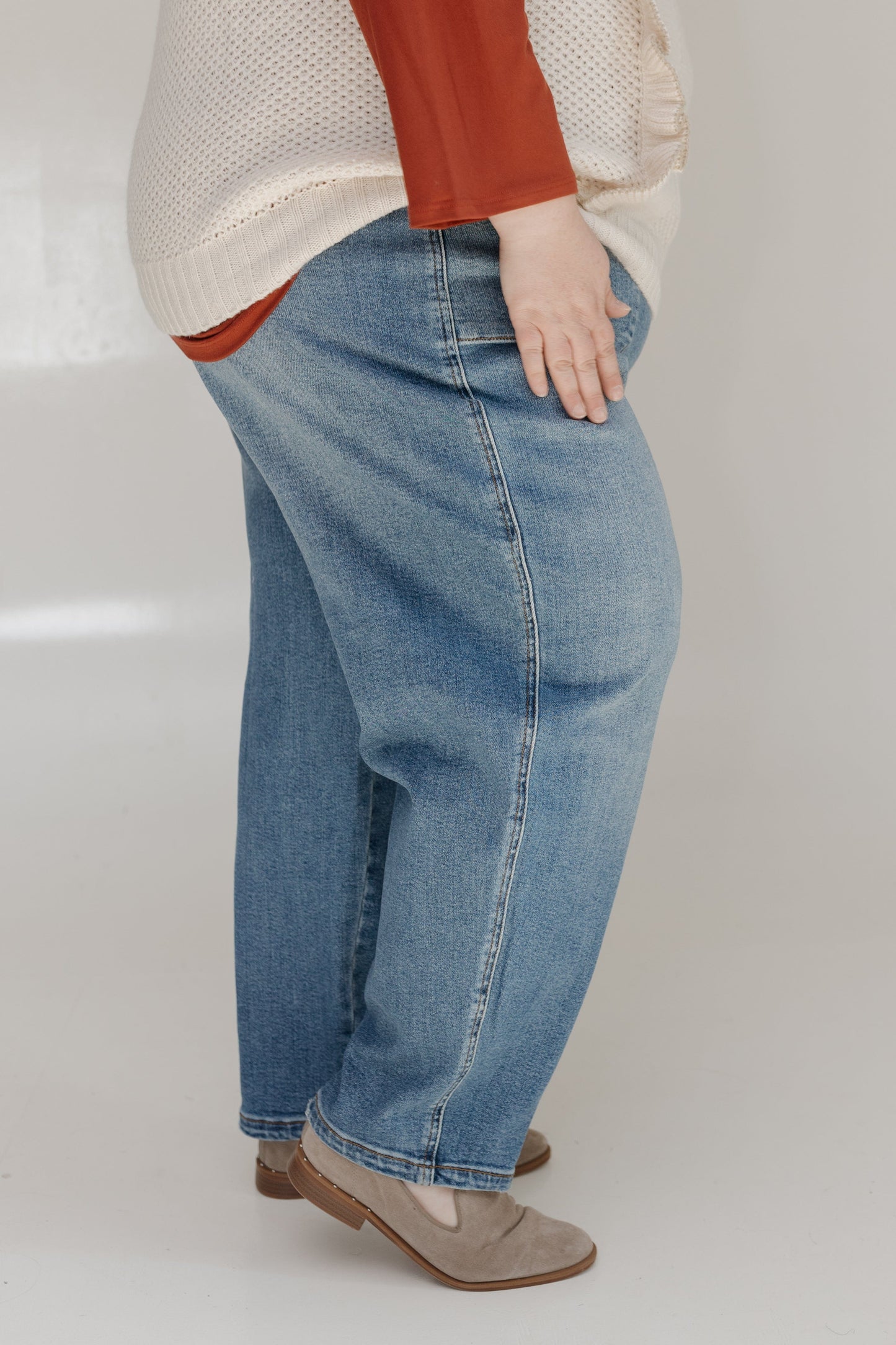 Person wearing blue jeans and a beige sweater with a red shirt underneath on a white background