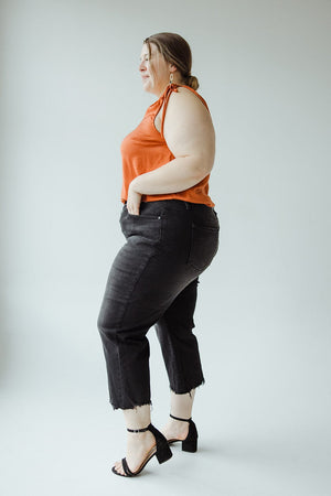 A woman stands against a plain background, wearing an orange sleeveless top, black cropped Judy Blue High Waist Button Fly Raw Hem Cropped Wide Leg jeans, and black strappy heels. She has her left hand in her pocket and is looking to her left.