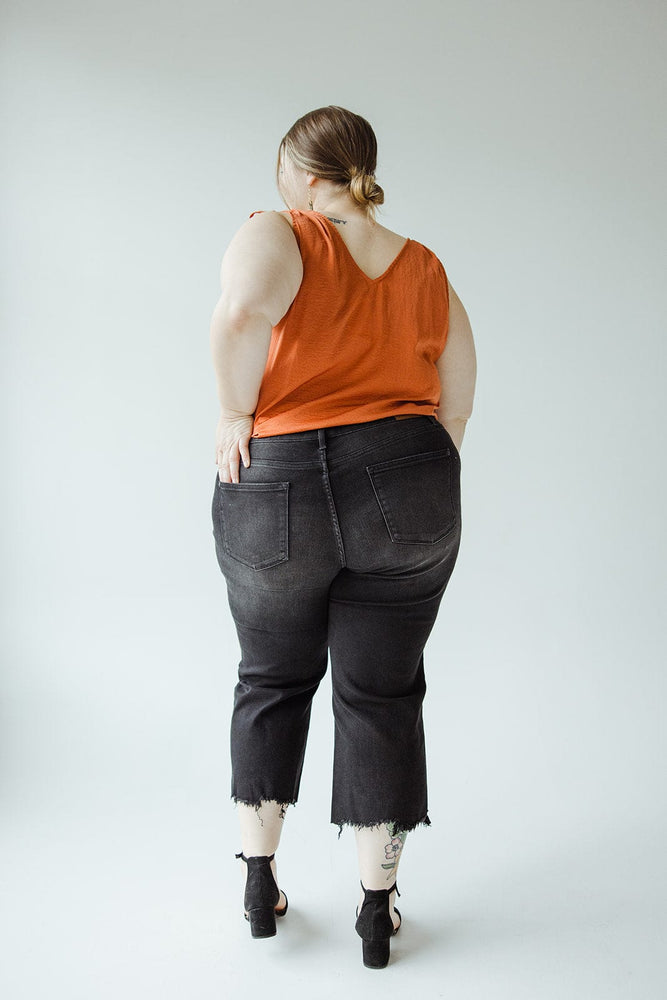 A person stands facing away, wearing a sleeveless orange top and high-waisted Judy Blue JUDY BLUE HIGH WAIST BUTTON FLY RAW HEM CROPPED WIDE LEG jeans. They have short hair and are positioned against a plain background.