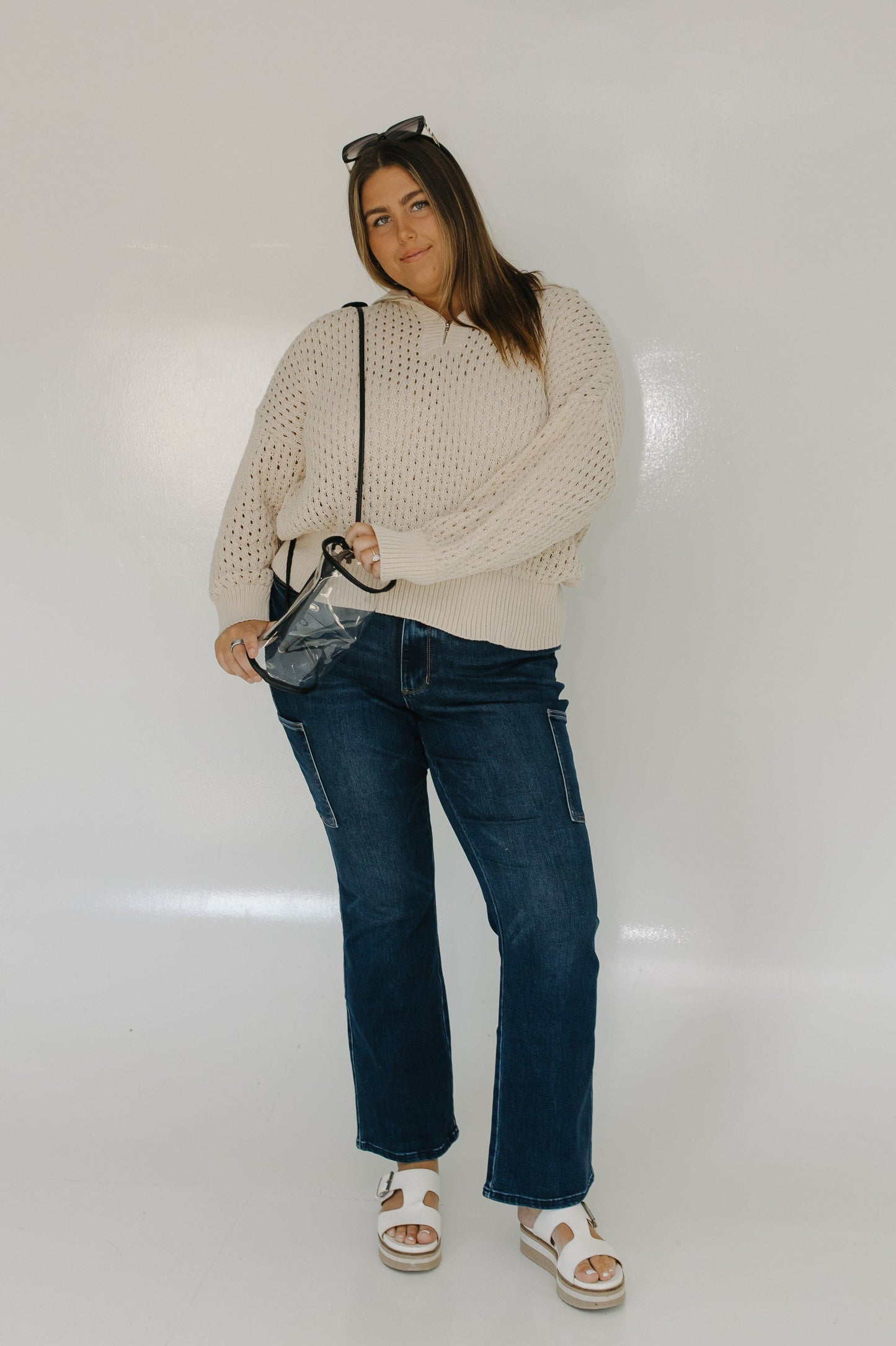 Woman wearing a beige sweater and blue jeans holding a clear handbag against a white background