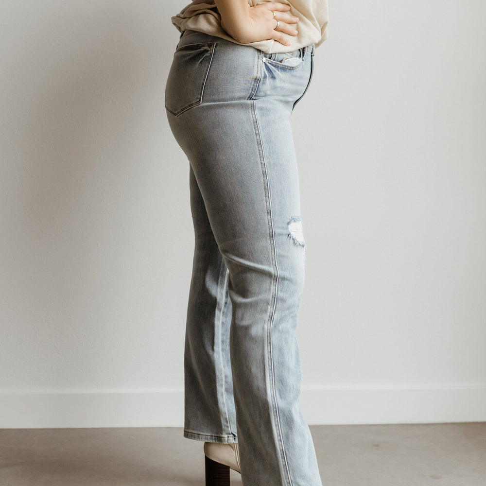 Person sporting a beige t-shirt, high-waist light blue jeans, and white heeled boots stands confidently with hands on hips against a plain backdrop.