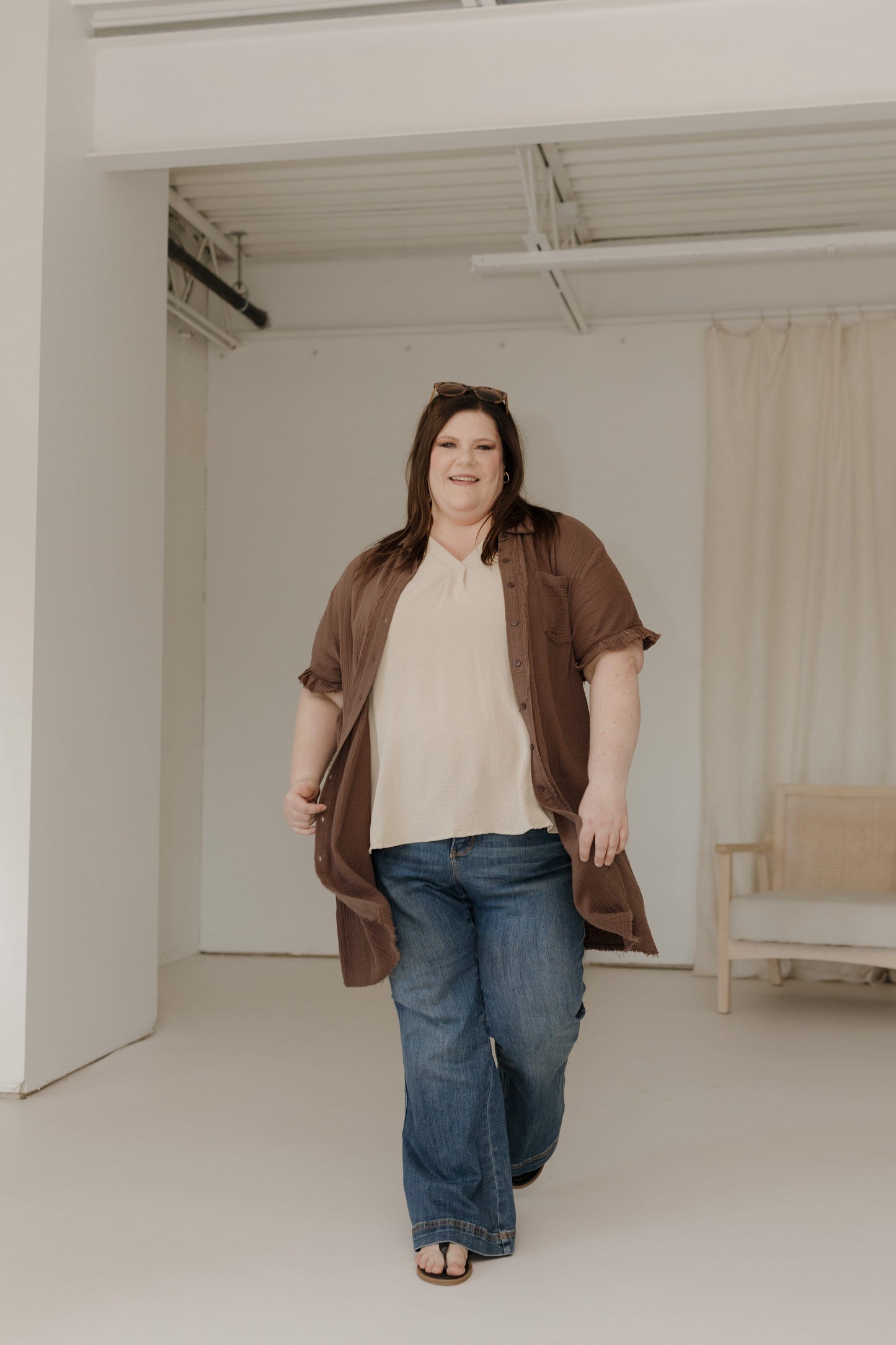 Woman wearing a brown jacket, beige top, and blue jeans in a minimalistic room.