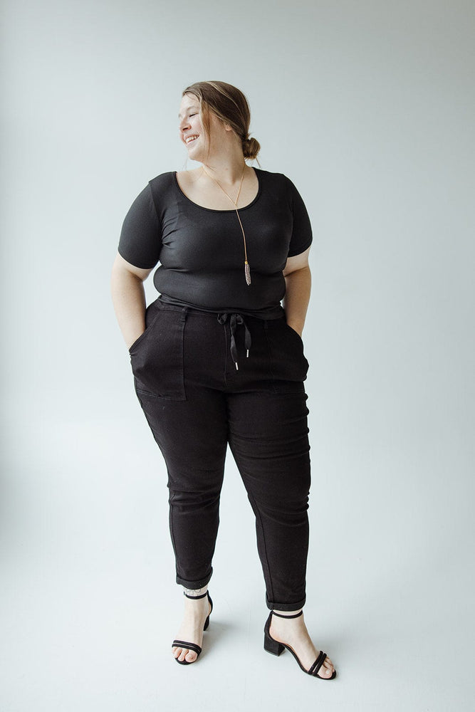 A person stands against a plain background, wearing Judy Blue JUDY BLUE HIGH WAIST DOUBLE ROLLED CUFFED JOGGER JEANS and black sandals, with hands in pockets and smiling.