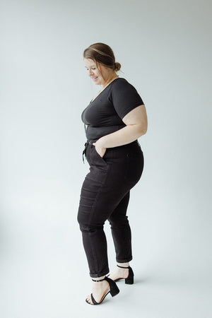 A woman stands sideways, looking down, wearing a black top, JUDY BLUE HIGH WAIST DOUBLE ROLLED CUFFED JOGGER JEANS for a perfect Judy Blue fit and black heels against a plain white background.