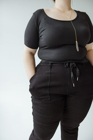 Person wearing a black short-sleeve top, high-waisted black pants, and a long necklace with a pendant, standing with hands in pockets against a plain background. The outfit includes JUDY BLUE HIGH WAIST DOUBLE ROLLED CUFFED JOGGER JEANS by Judy Blue that's perfect for any occasion.