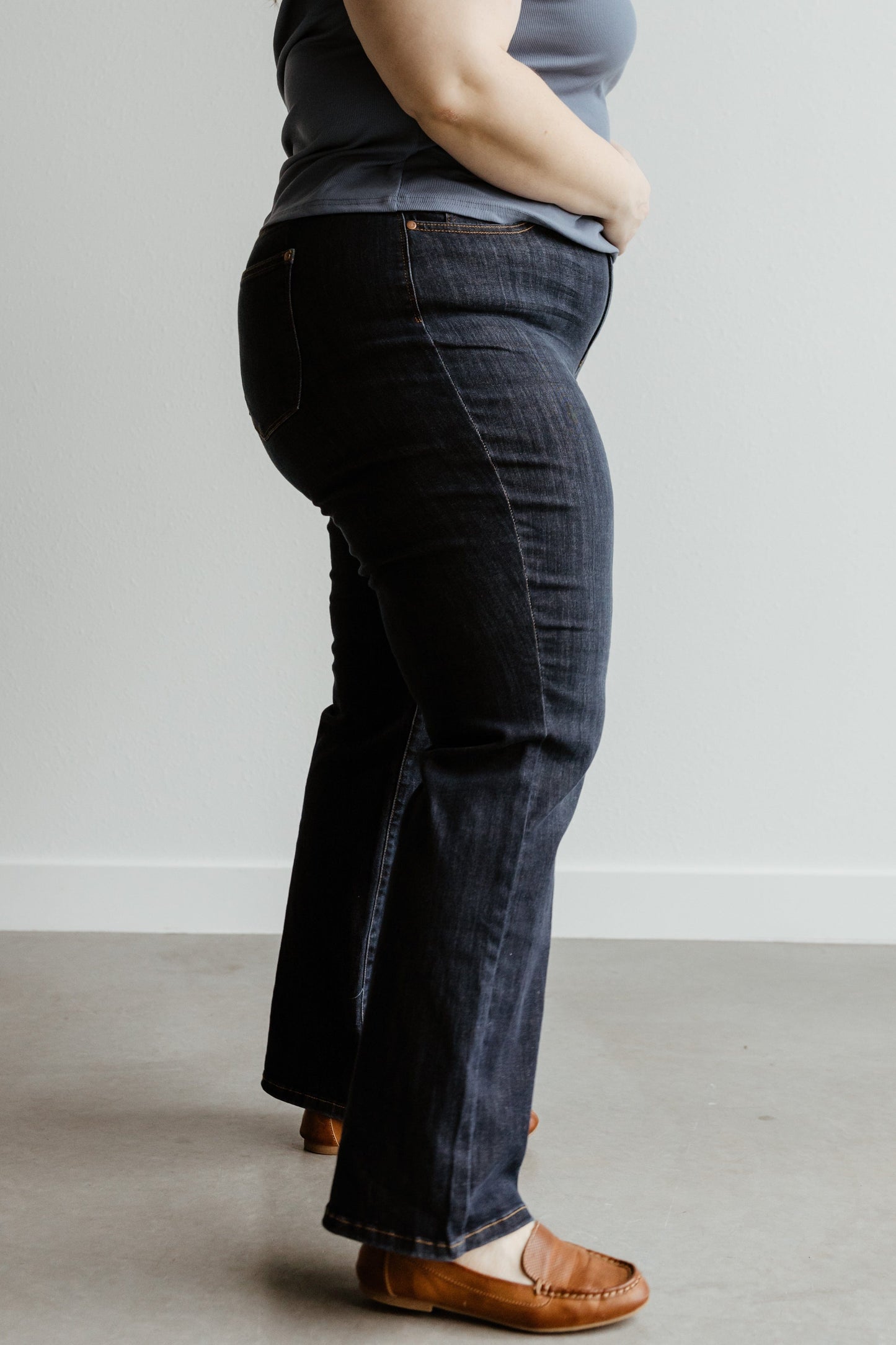 A person wearing a grey top, Judy Blue Jeans, and brown shoes stands side-on against a plain background.