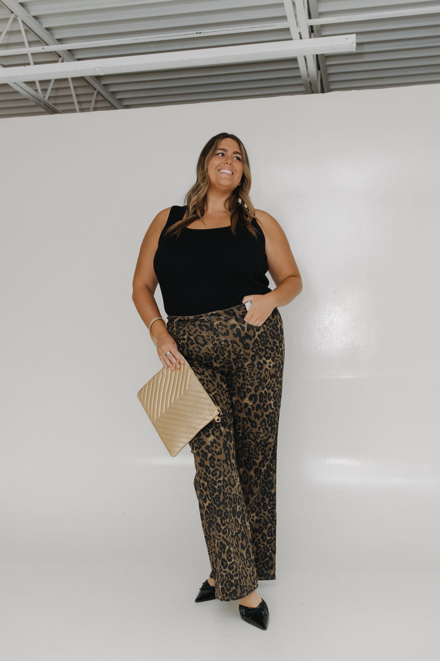 Woman wearing a black top and leopard print pants with a beige clutch against a white wall.