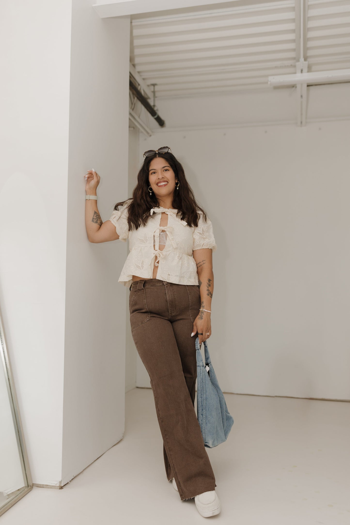 Woman in a white blouse and brown pants standing in a room with a mirror.