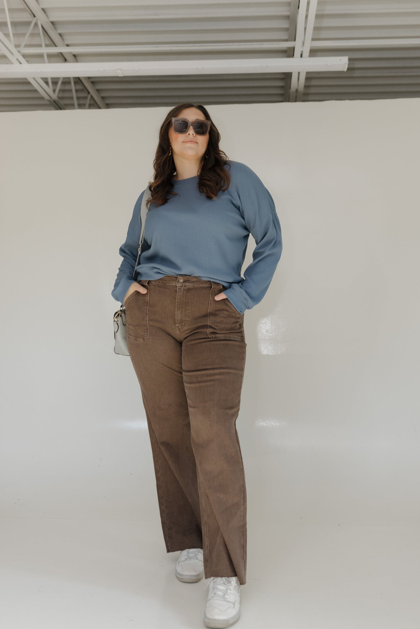 Person wearing a blue sweater, brown pants, and white sneakers in a minimal indoor setting.