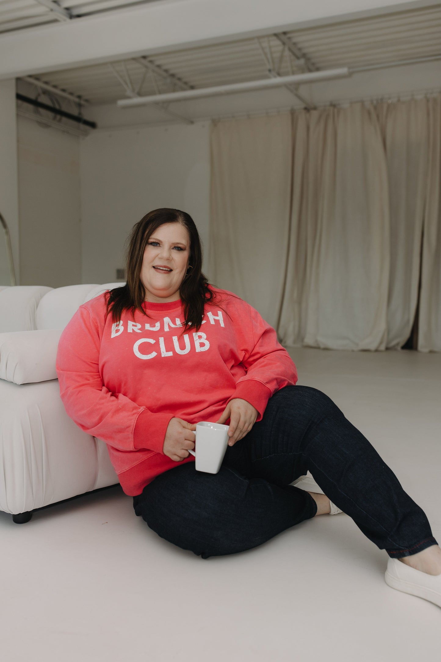 Person wearing a pink 'Brunch Club' sweatshirt sitting on a white couch holding a white mug.