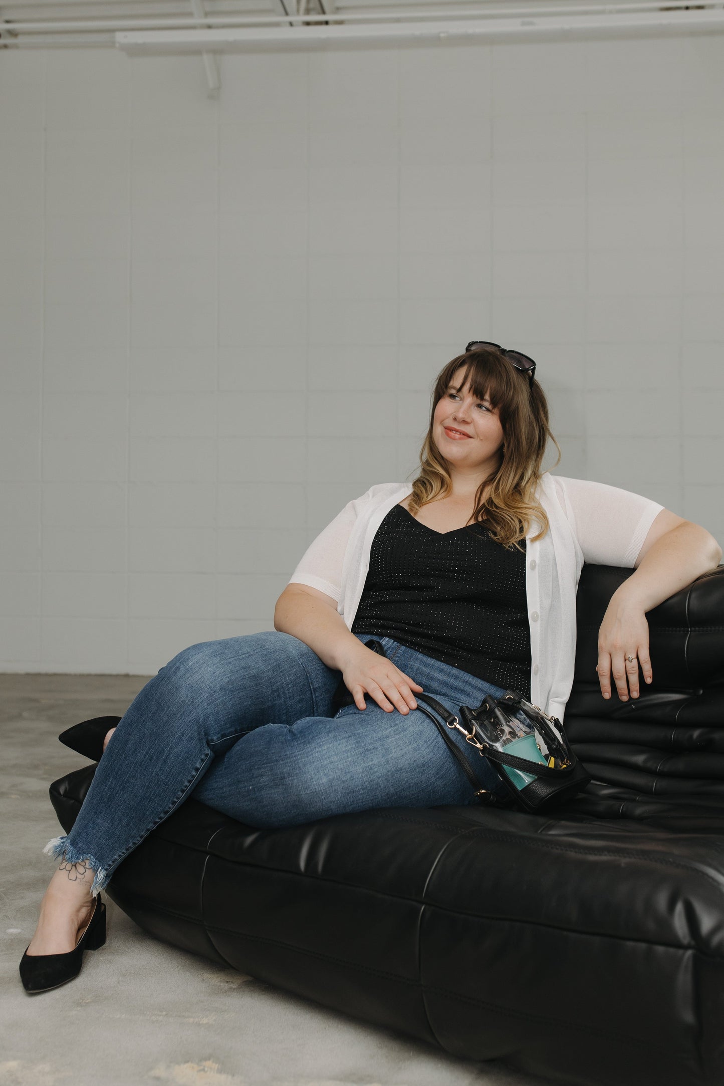 Woman sitting on a black leather couch wearing a white cardigan, black top, and blue jeans.