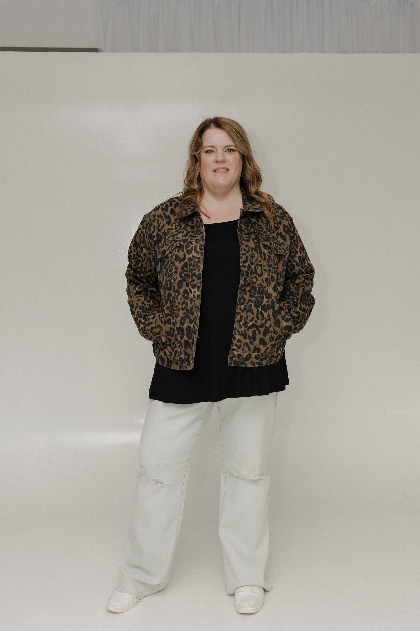 Person wearing a leopard print jacket, black top, white pants, and white shoes against a plain background
