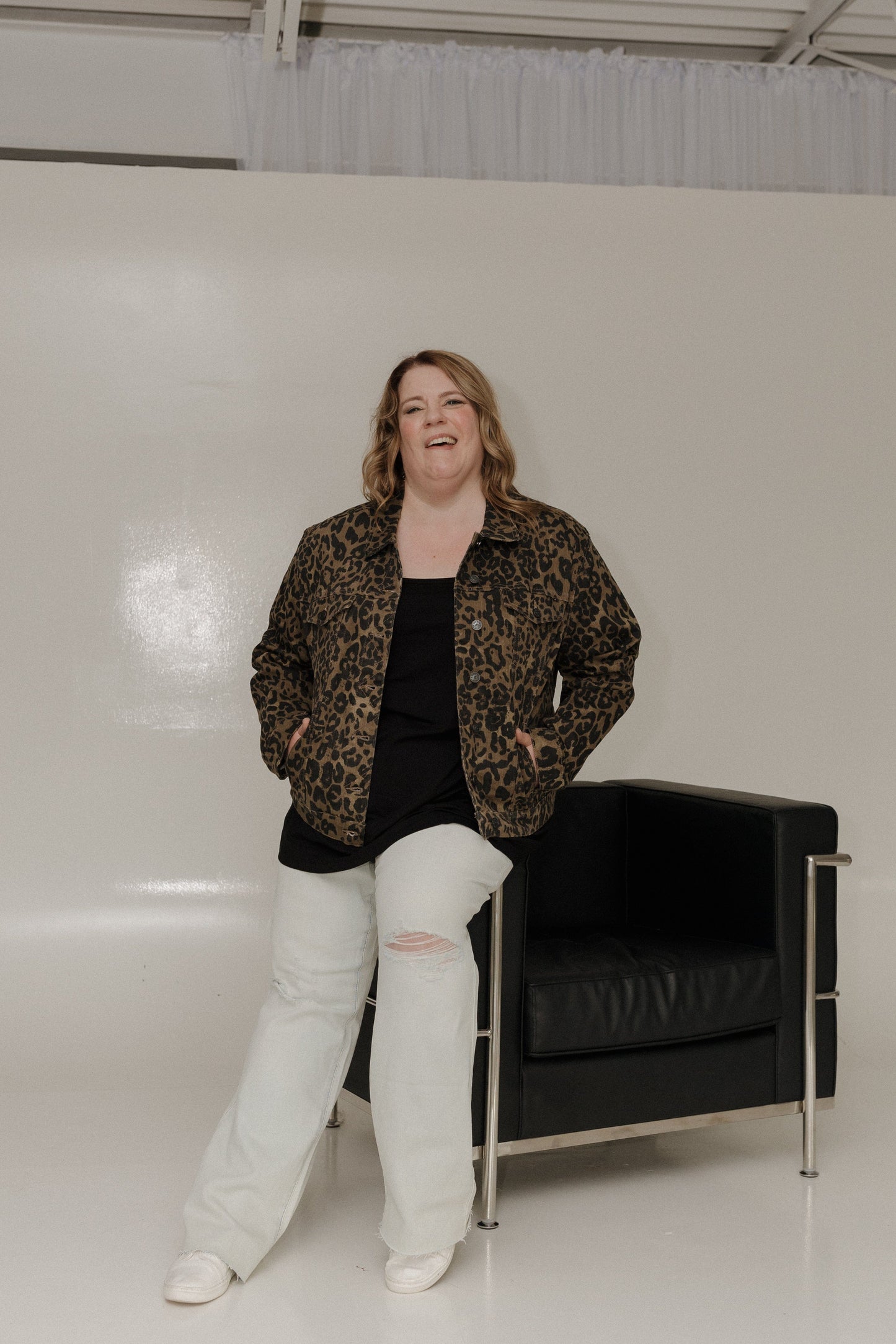 Person wearing a leopard print jacket and white pants standing next to a black chair against a plain background