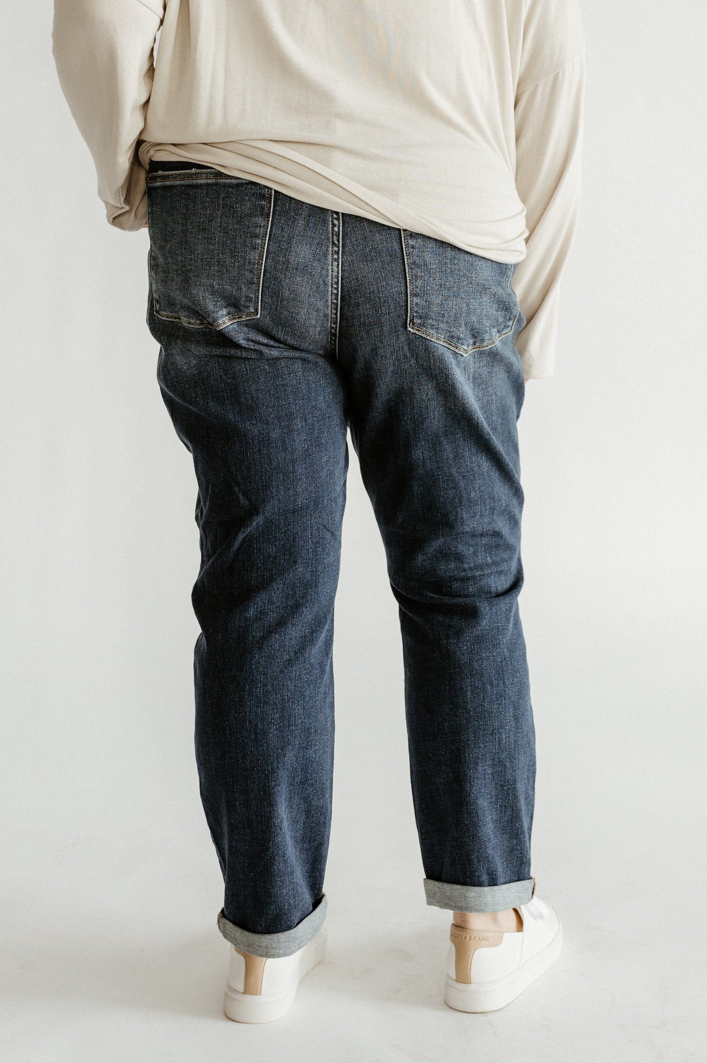 A person wearing a light shirt, MID-RISE CAT SCRATCH BOYFRIEND JEANS, and white sneakers stands with their back facing the camera.