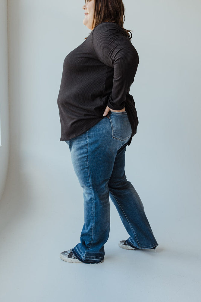 A person with long hair, wearing a black top and blue JUDY BLUE MID-RISE DAD JEAN WITH RELEASED HEM by Judy Blue, is standing with their back to the camera, hands resting on their hips.