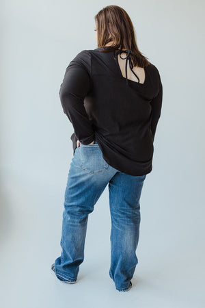 A person with long hair, wearing a black top and blue JUDY BLUE MID-RISE DAD JEAN WITH RELEASED HEM by Judy Blue, is standing with their back to the camera, hands resting on their hips.