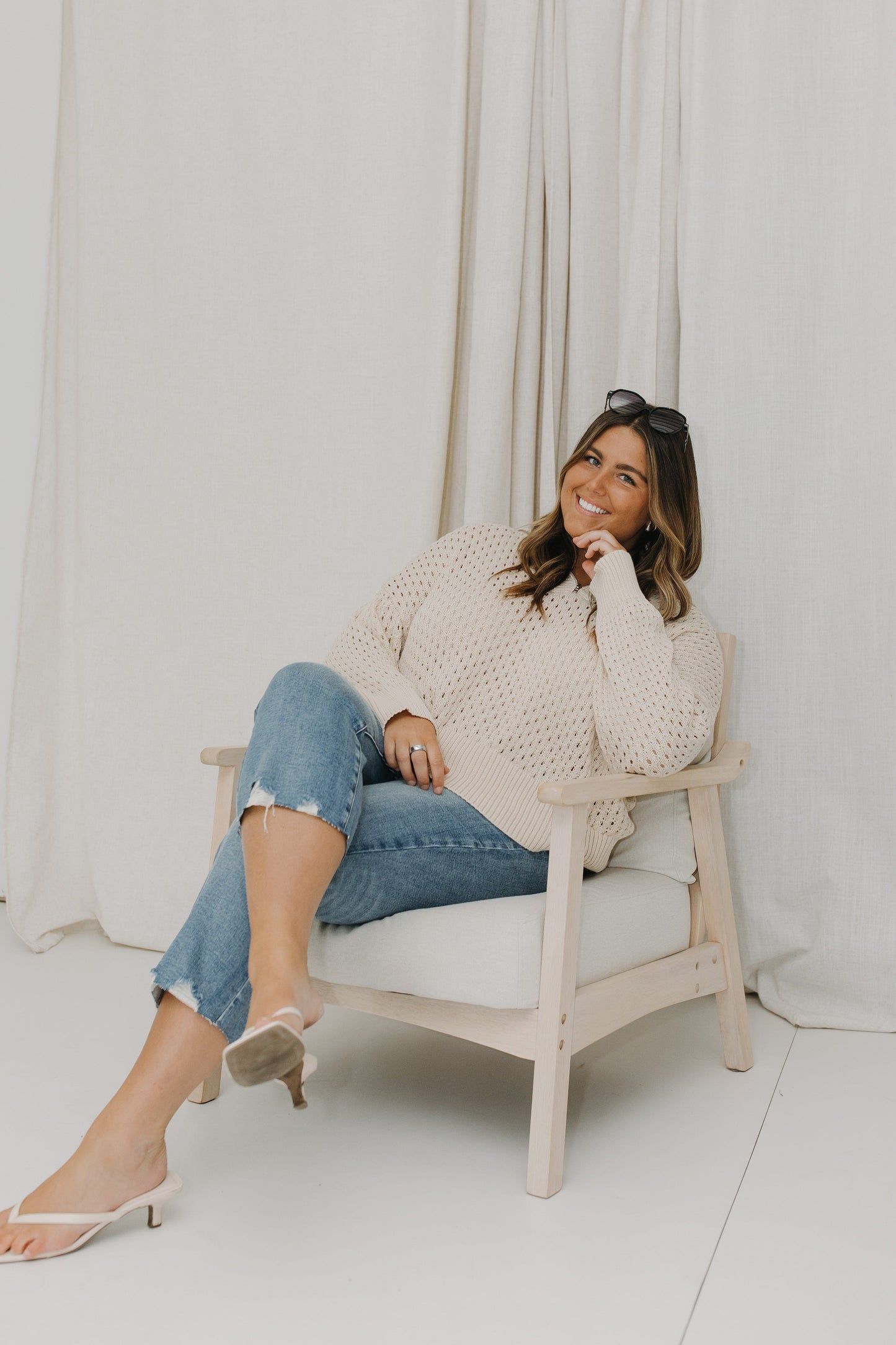 Woman sitting on a chair wearing a white sweater and blue jeans against a white curtain background