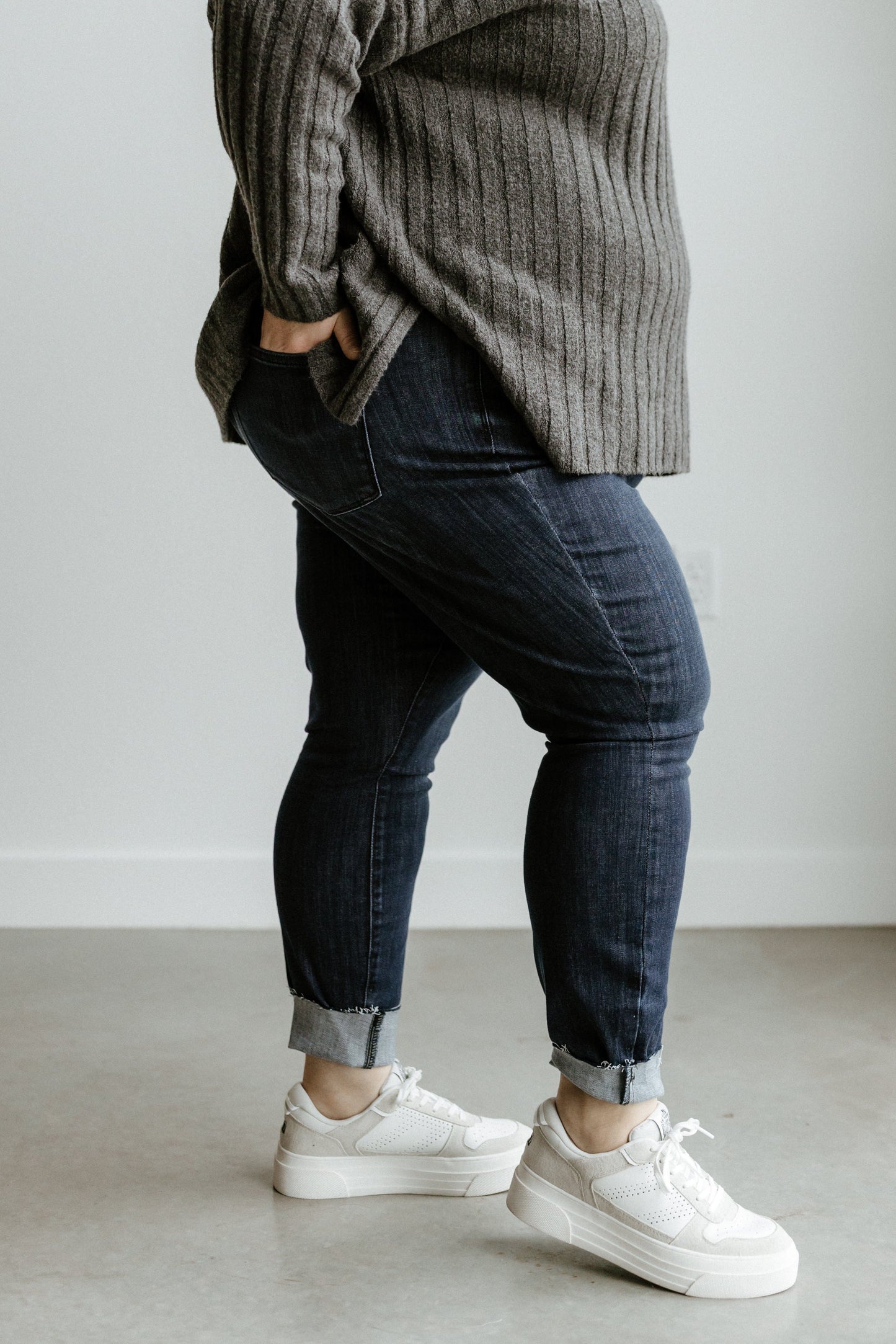 A person wearing a gray sweater, Judy Blue mid-rise skinny jeans, and white sneakers stands indoors on a light-colored floor.