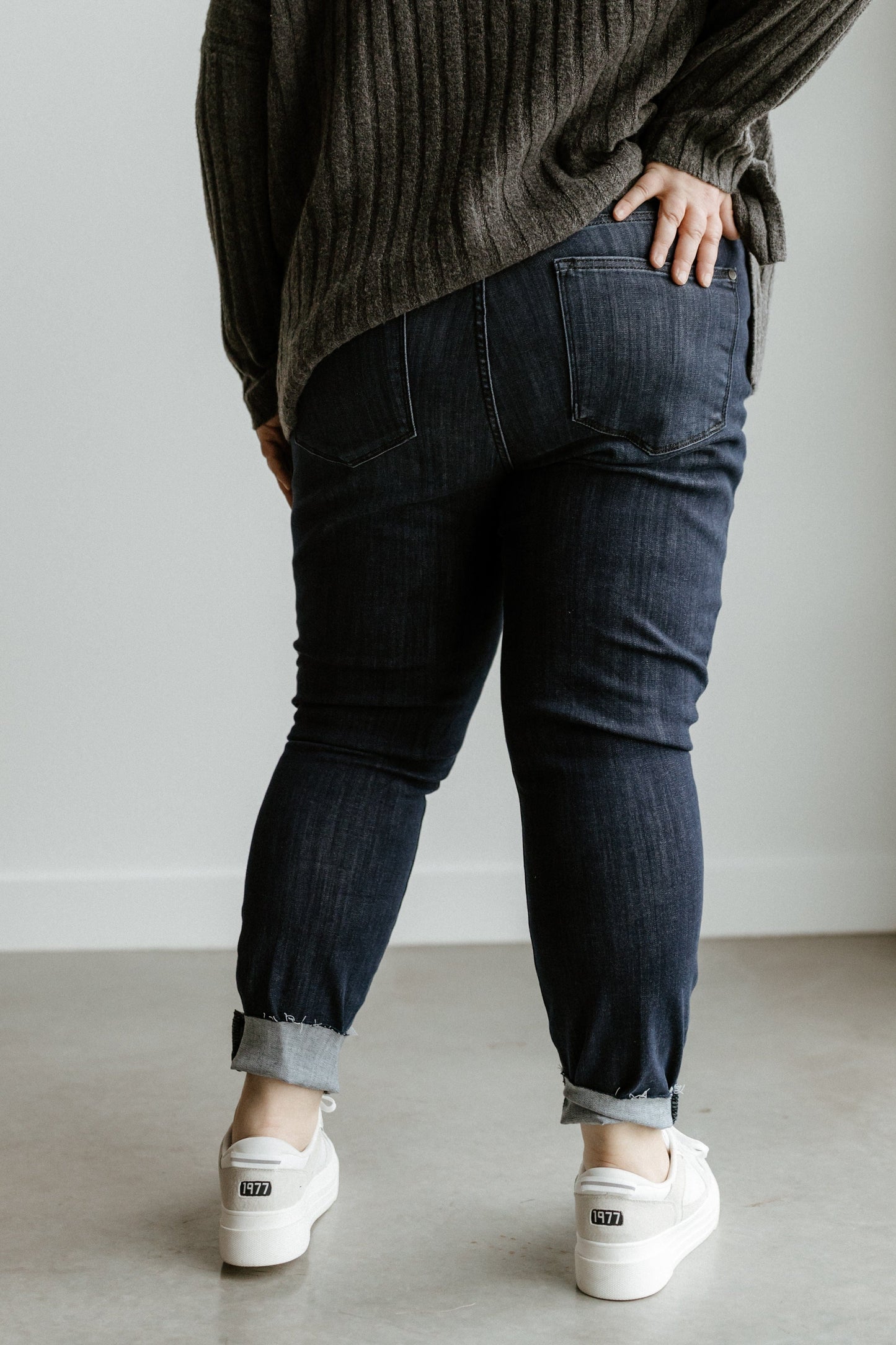 A person in Judy Blue mid-rise skinny jeans and a gray sweater stands against a white wall, seen from behind.