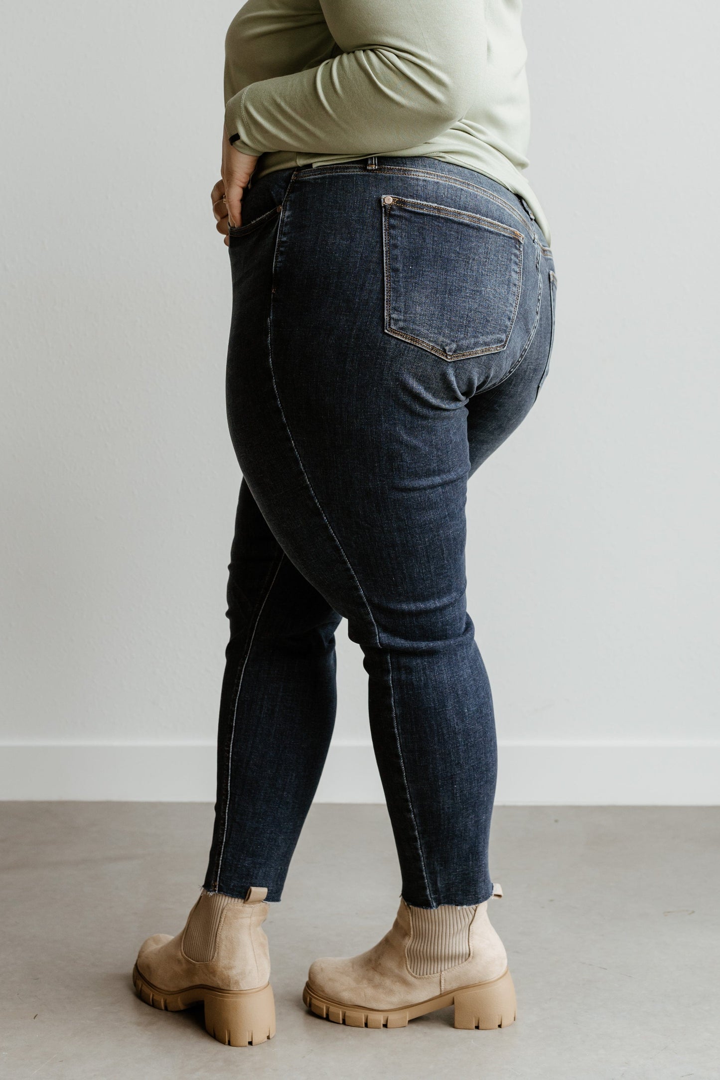 A person wearing a light green top, beige boots, and mid-rise skinny jeans stands sideways in front of a plain white wall.