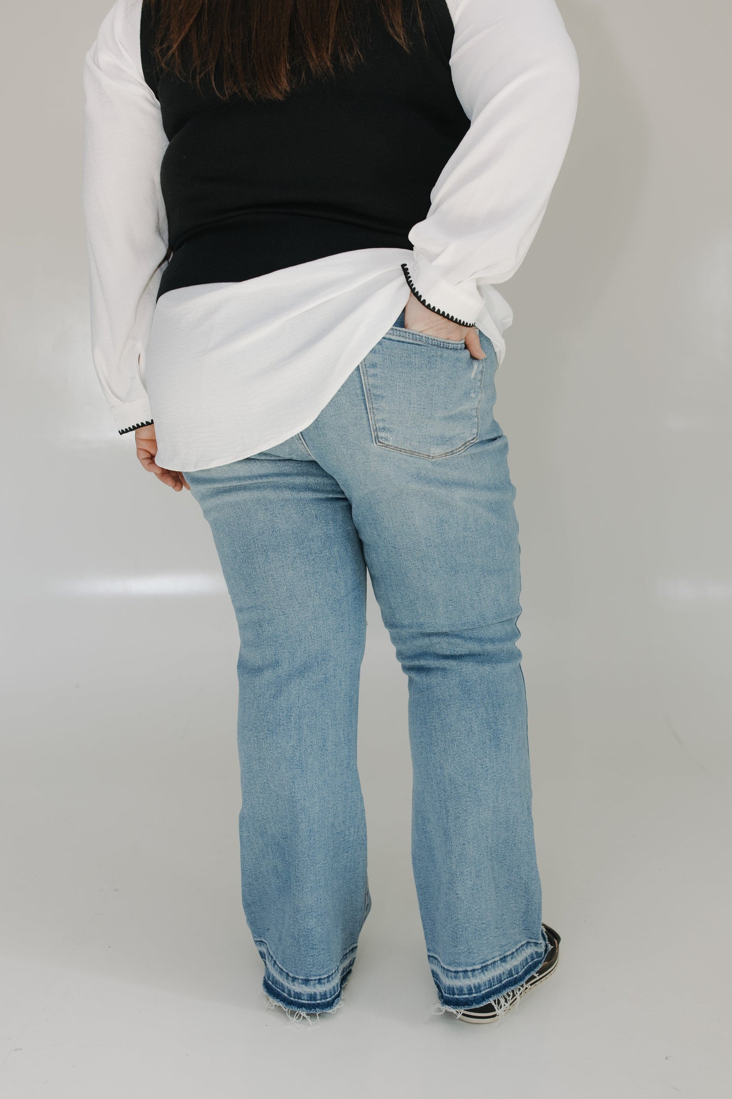 Person wearing a white long-sleeve shirt, black vest, and light blue jeans on a plain background