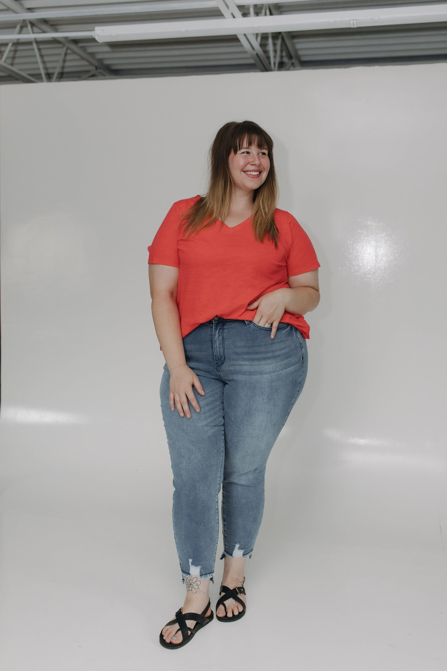 Woman wearing a red shirt and blue jeans standing against a white wall.