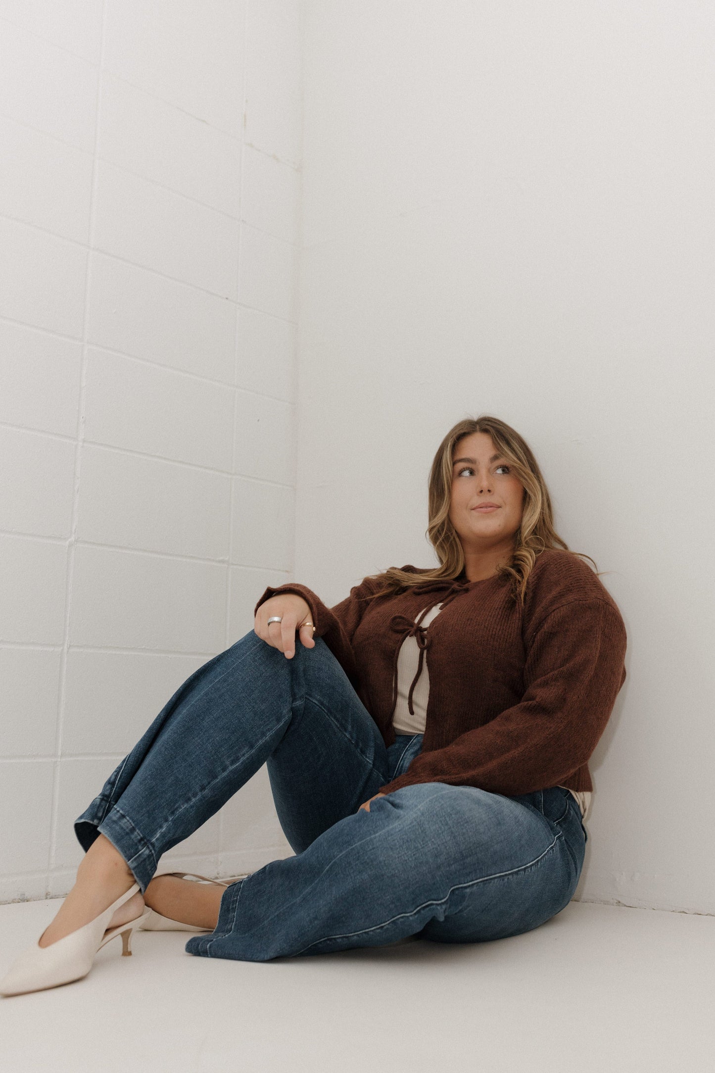 Woman sitting against a white brick wall wearing a brown sweater and blue jeans.
