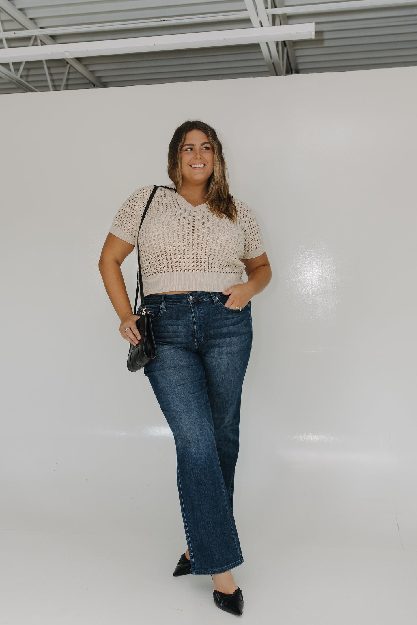 Woman wearing a beige top and blue jeans standing against a white wall.