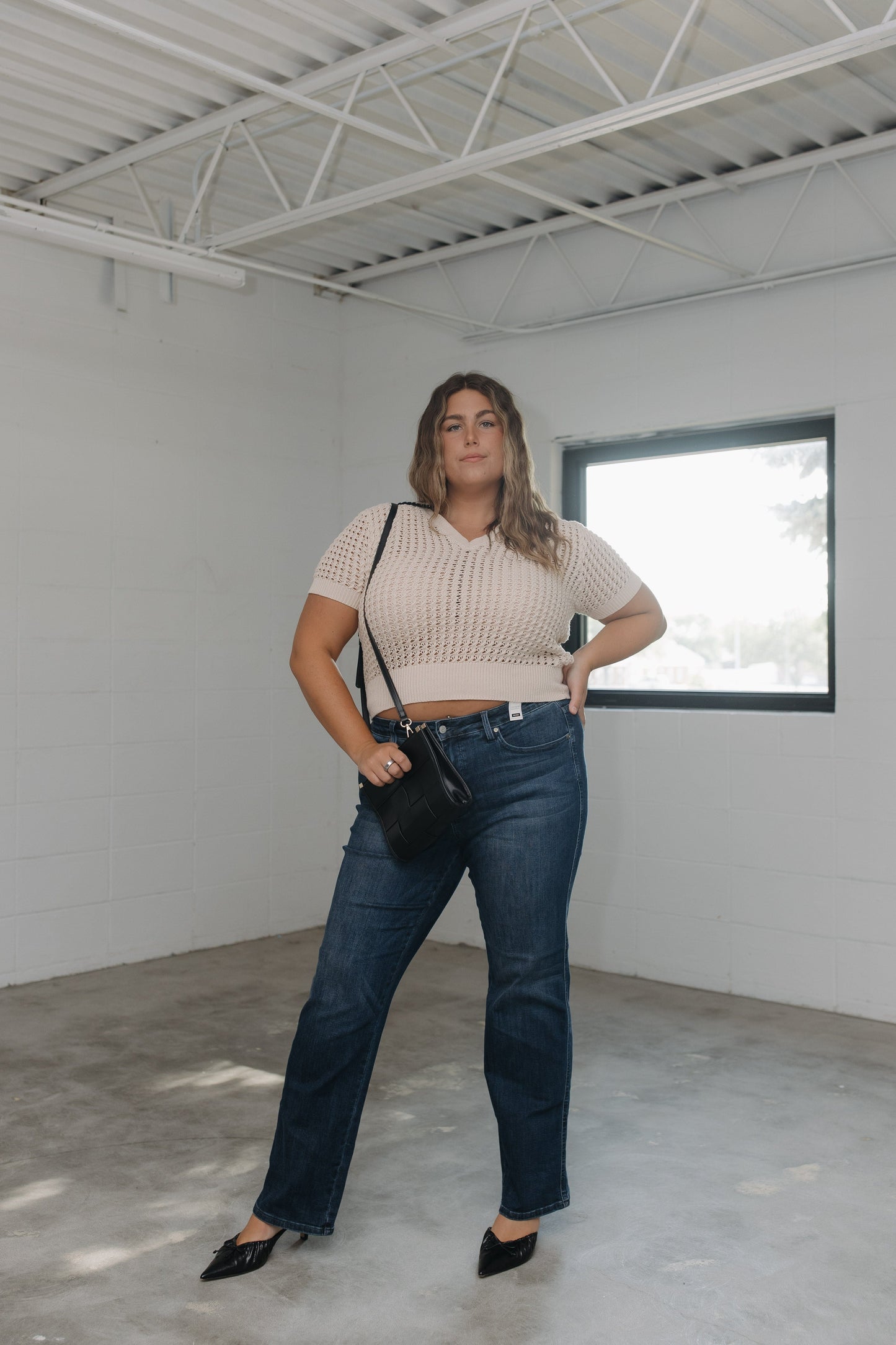 Woman in a beige crop top and blue jeans standing in a minimalistic room with a window.