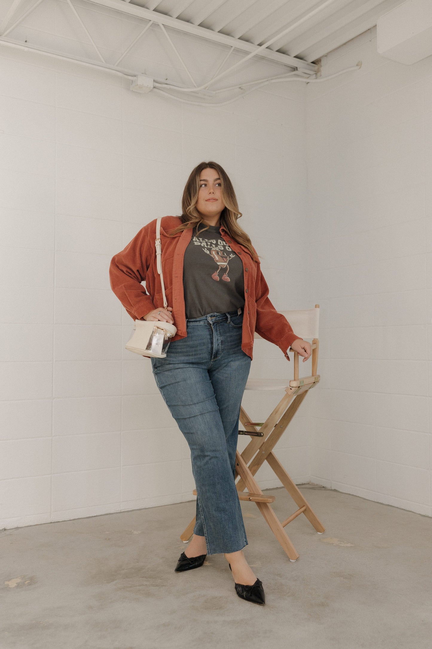 Woman in a red jacket and blue jeans standing in a minimalistic room with a chair and bag.