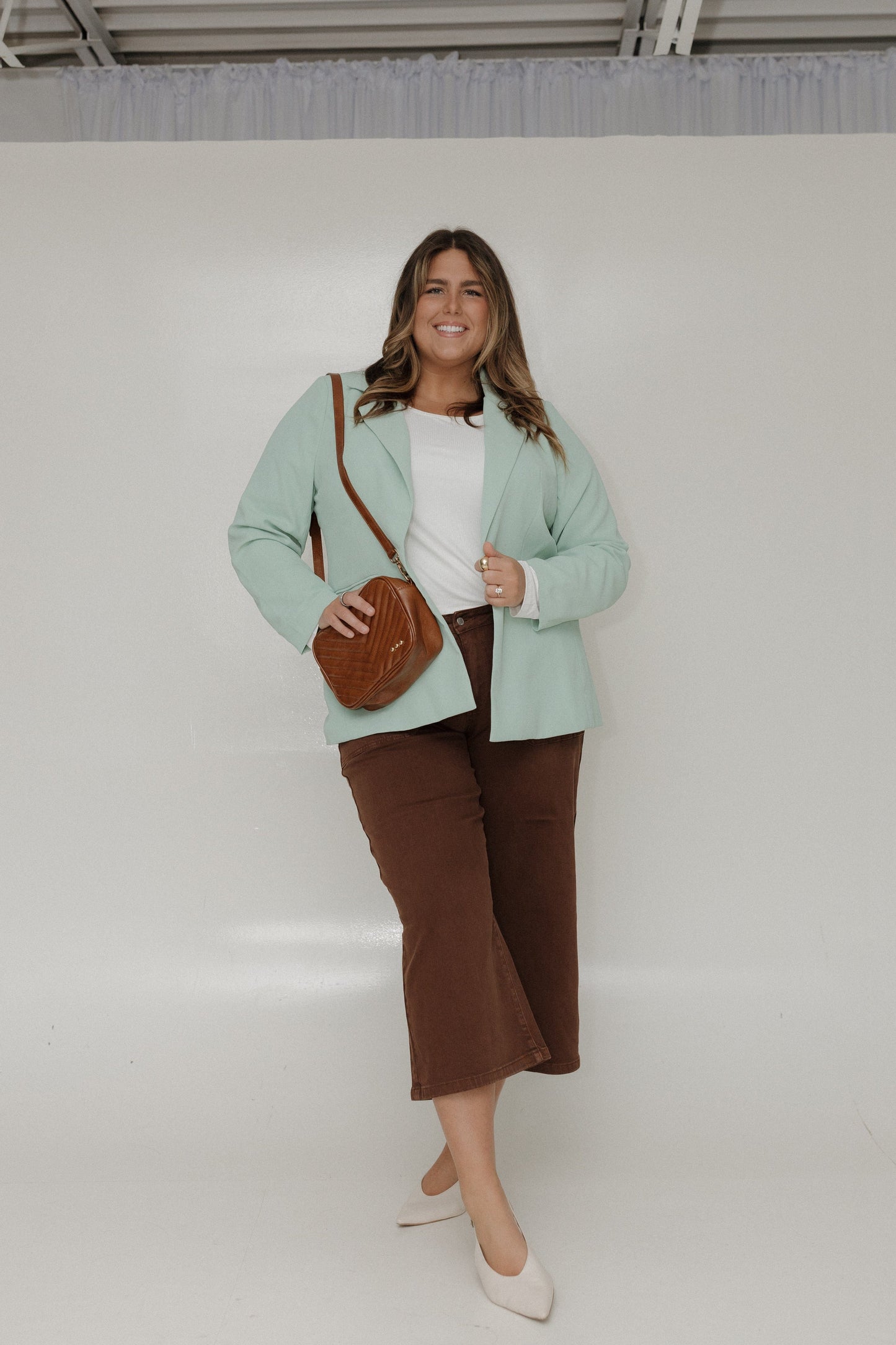 Woman wearing a light green blazer and brown pants, holding a brown handbag against a white background