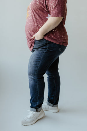 A person wearing a maroon shirt, Judy Blue Jeans, and white sneakers stands with one hand in their pocket.