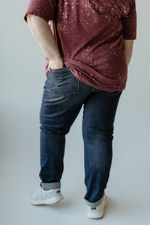 A person wearing a maroon speckled shirt, cuffed slim fit jeans, and white sneakers stands with one hand in a back pocket, facing away from the camera.